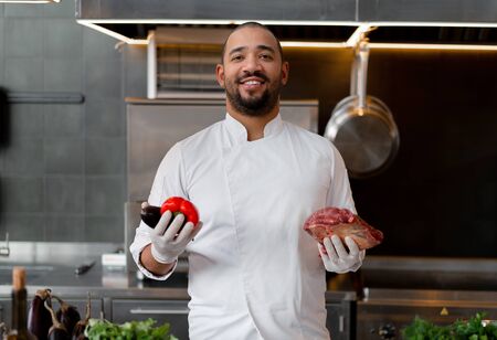 Happy smiling chef prepares meat dish with various vegetables in the kitchen. In one hand the man holds vegetables, in the other a piece of fresh meat.の写真素材