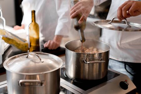 Closeup hands of unrecognizable chef pouring wine into pot with meat. In the background is a blurred figure of another chef.の写真素材