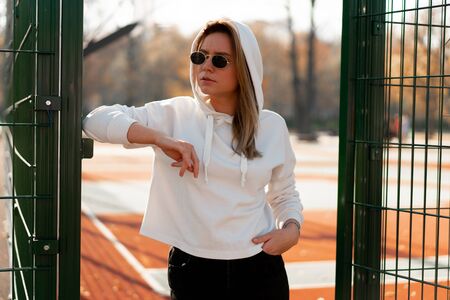 Outdoor close up portrait of young beautiful woman with long hair in sunglasses, dressed in a white hoodie sweater, near the sportsground. youth culture summer pastimeの写真素材