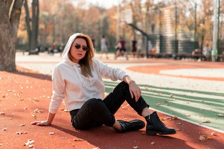 Outdoor portrait of young beautiful woman with long in sunglasses and a white hooded sweater sitting on the sportsground track.  youth culture summer pastimeの写真素材