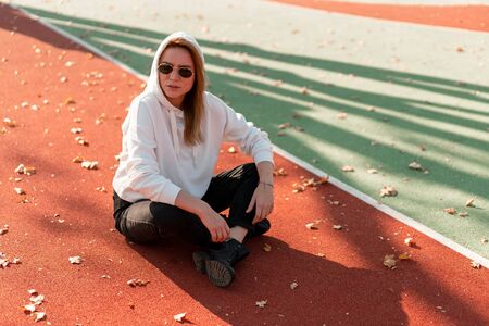 Outdoor portrait of young beautiful woman with long in sunglasses and a white hooded sweater sitting on the sportsground track.  youth culture summer pastimeの写真素材