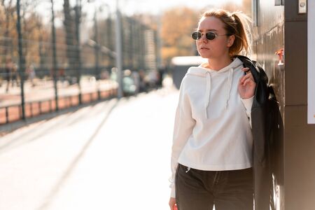 Outdoor portrait of young beautiful woman with a ponytail and sunglasses, with a backpack on his shoulders, dressed in a white sweater, near the sportsground. White hoodieの写真素材
