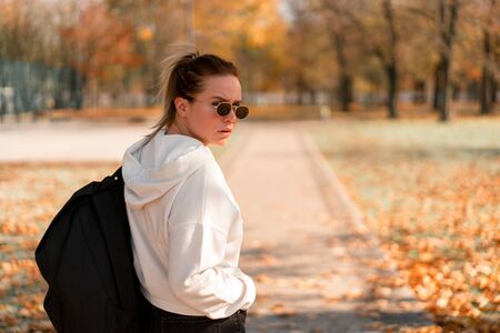 A young beautiful woman with a ponytail and sunglasses, with a backpack on his shoulders in the park. Photo from the back. White hoodieの写真素材