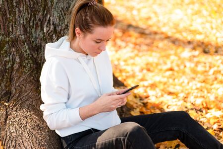 Woman in a white hoodie sweater with a hood sits on ground in the park and holding a cellphone in her hands. cute girl use smartphone for communication Female texting a message Communicaion conceptの写真素材