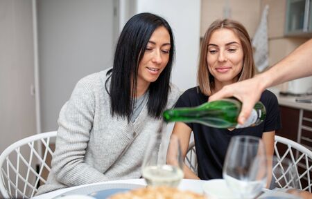 Friends meeting with wine and cake in the modern style kitchen. Young women smile and joke. A male hand in the foreground pours wine into glasses.の写真素材