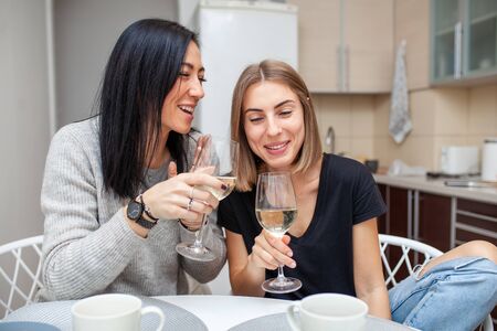 Friends meeting with wine and cake in the modern style kitchen. Young women smile and joke with glasses of wine in his hands. Two girls drink wine in the home kitchen.の写真素材