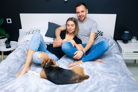 Young couple sitting on a bed in the bedroom, hugging. Near them is their dog. Domestic animal Beagleの写真素材