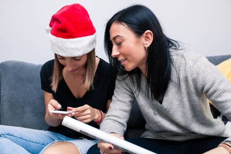 Two young women are preparing for the new year looking in mobile phone ways to pack gifts. New Year celebration. Two girlfriends at the New Year holiday read SMS on a mobile phone.の写真素材