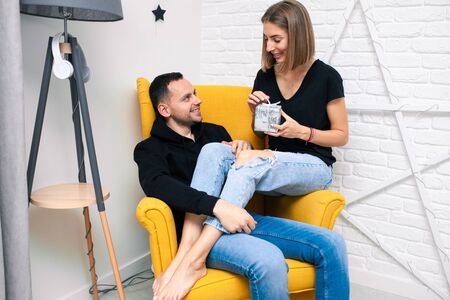 Portrait of couple young in a cozy room. A man sits on a yellow chair. A woman sits on the lap of a man and holds a gift Christmas family holidays. Modern style interior.の写真素材