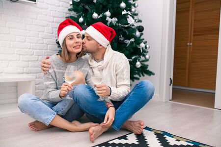 Young happy couple in Christmas hats near a Christmas tree kissing, holding glasses of wine. New Year celebration. Happy New Year Family Concept. Christmas for twoの写真素材