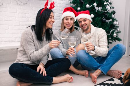 Happy friends celebrating New Year in home interior in Christmas hats sitting near a Christmas tree with glasses of wine.の写真素材