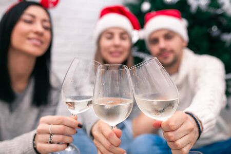 Happy friends celebrating New Year in home interior in Christmas hats sitting near a Christmas tree with glasses of wine. Wine glasses are the focus. Blurred figures of people behind glasses.の写真素材