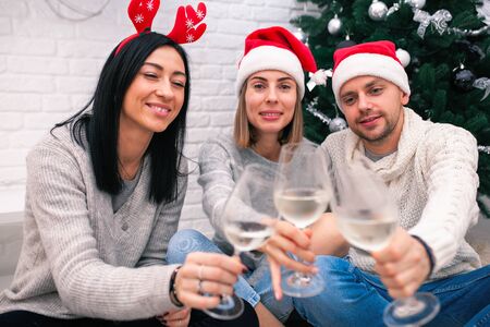 Happy friends celebrating New Year in home interior in Christmas hats sitting near a Christmas tree with glasses of wine.の写真素材