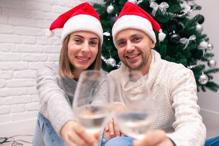 Young happy couple in Christmas hats near a Christmas tree and holding glasses of wine. New Year celebration.の写真素材