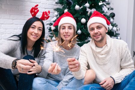 Happy friends celebrating New Year in home interior in Christmas hats sitting near a Christmas tree with sparkler. Bengal light are the defocusedの写真素材