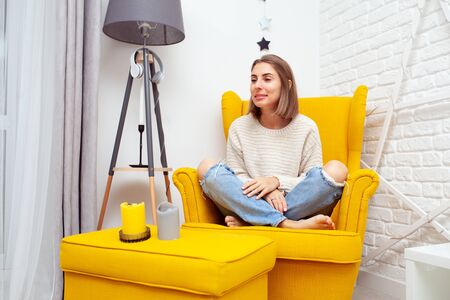 Enjoying leisure time at home. Beautiful young woman sitting in a soft yellow chair resting. Aromatic candles stand on the side.の写真素材