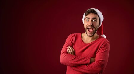 Young handsome caucasian guy in red sweater and Santa hats stands on red background in studio and teeth smiling. Close up portrait european guy with Christmas mood. He folded his arms over his chest.の写真素材