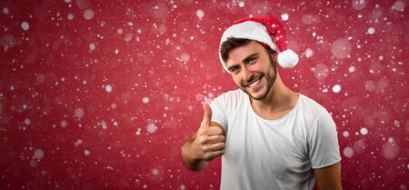 Young handsome caucasian guy in white t-shirt and Santa hats stands on red background in studio showing thumbs up. Close up portrait european guy with Christmas mood with snow Holiday banner.の写真素材
