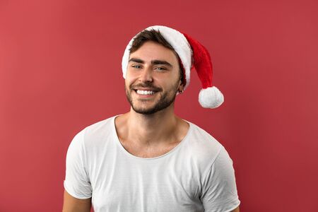 Young handsome caucasian guy in a white t-shirt and Santa hats stands on red background in studio and teeth smiling. Close up portrait european guy with Christmas mood. Holiday theme. New Year.の写真素材