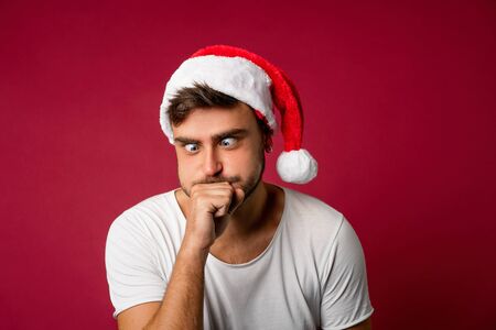 Young handsome caucasian guy in a white t-shirt and Santa hats stands on red background in studio and Sick after holiday food. Close up portrait european guy with Christmas mood. Holiday theme. Jokingの写真素材