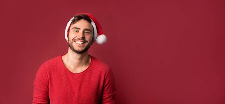Young handsome caucasian guy in red sweater and Santa hats stands on red background in studio and teeth smiling. Close up portrait european guy with Christmas mood. Holiday banner. New Year.の写真素材
