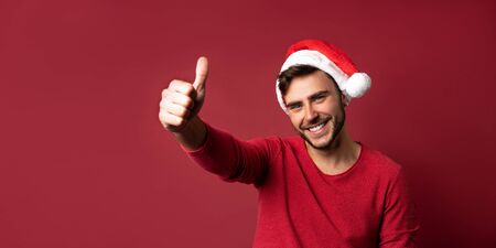 Young handsome caucasian guy in red sweater and Santa hats stands on red background in studio and showing thumbs up. Close up portrait european guy with Christmas mood. Holiday banner.の写真素材