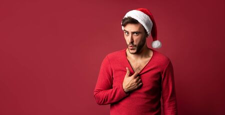 Young handsome caucasian guy in Santa hats stands on red background in studio pulls the sweater Shows how hot he is. Close up portrait european guy with Christmas mood. Holiday banner. New Year.の写真素材