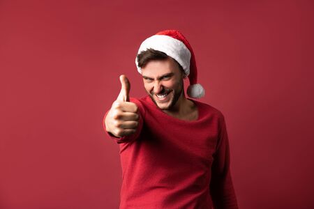 Young handsome caucasian guy in red sweater and Santa hats stands on red background in studio and showing thumbs up. Close up portrait european guy with Christmas demonic mood. Holiday banner.の写真素材