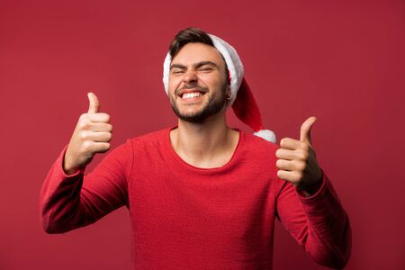 Young handsome caucasian guy in red sweater and Santa hats stands on red background in studio and showing thumbs up with two hand. Close up portrait european guy with Christmas mood. Holiday theme. New Year.の写真素材