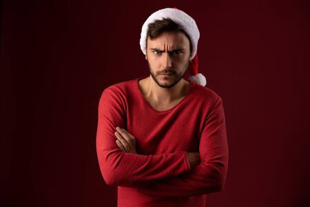 Young handsome caucasian guy in red sweater and Santa hats stands on red background in studio and feel angry. Close up portrait european guy with Christmas mood. Holiday theme. New Year.の写真素材