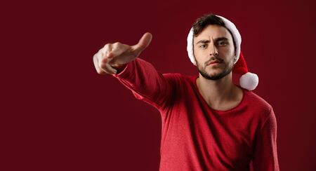 Young handsome caucasian guy in red sweater and Santa hats stands on red background in studio and Shows finger at the camera Close up portrait european guy with Christmas mood Holiday banner New Yearの写真素材