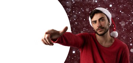 Young handsome caucasian guy in red sweater and Santa hats stands on red background in studio and Shows finger at the camera Close up portrait european guy with Christmas mood Holiday banner New Yearの写真素材