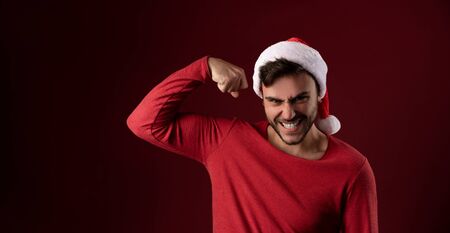 Young handsome caucasian guy in red sweater and Santa hats stands on red background in studio and shows biceps power. Close up portrait european guy with Christmas mood. Holiday banner. New Year.の写真素材