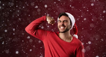 Young handsome caucasian guy in red sweater and Santa hats stands on red background in studio and shows biceps power. Close up portrait european guy with Christmas mood. Holiday banner. New Year.の写真素材