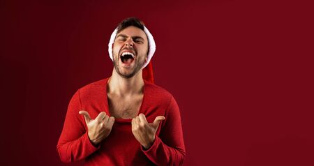 Young handsome caucasian guy in Santa hats stands on red background in studio pulls the sweater Shows how hot he is. Close up portrait european guy with Christmas mood. Holiday banner. New Year.の写真素材