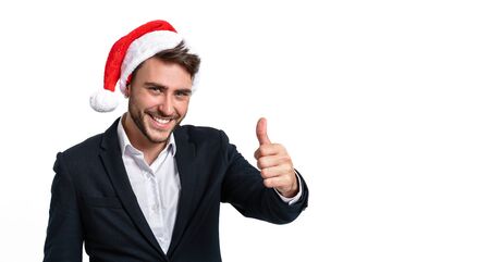 Young handsome caucasian guy in business suit and Santa hats stands on white background in studio smilie and showing thumbs up. Close up portrait business person with Christmas mood Holiday bannerの写真素材