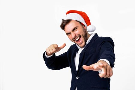 Young handsome caucasian guy in business suit and Santa hats on white background in studio smilie and showing Finger to the camera. Close up portrait business person with Christmas mood Holiday bannerの写真素材