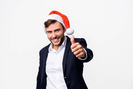 Young handsome caucasian guy in business suit and Santa hats stands on white background in studio smilie and showing thumbs up. Close up portrait business person with Christmas mood Holiday bannerの写真素材