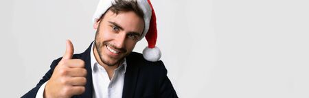 Young handsome caucasian guy in business suit and Santa hats stands on white background in studio smilie and showing thumbs up. Close up portrait business person with Christmas mood Holiday bannerの写真素材