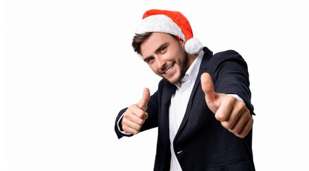 Young handsome caucasian guy in business suit and Santa hats on white background in studio smilie and showing thumbs up two hands. Close up portrait business person with Christmas mood Holiday bannerの写真素材