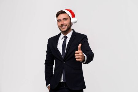 Young handsome caucasian guy in business suit and Santa hats on white background in studio smilie and showing thumbs up two hands. Close up portrait business person with Christmas mood Holiday bannerの写真素材