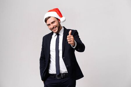 Young handsome caucasian guy in business suit and Santa hats on white background in studio smilie and showing thumbs up two hands. Close up portrait business person with Christmas mood Holiday bannerの写真素材
