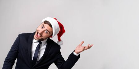 Young handsome caucasian guy in business suit and Santa hats on white background in studio smilie and showing hand to the side. Close up portrait business person with Christmas mood Holiday bannerの写真素材