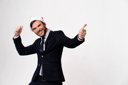 Young handsome caucasian guy in business suit and Santa hats on white background in studio smilie and showing thumbs up two hands. Close up portrait business person with Christmas mood Holiday bannerの写真素材