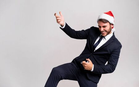 Young handsome caucasian guy in business suit and Santa hats stands on white background in studio Plays on an imaginary guitar. Close up portrait business person with Christmas mood Holiday bannerの写真素材