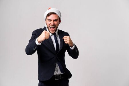 Young handsome caucasian guy in business suit and Santa hats on white background studio screams and fights hold fists for sparring Close up portrait business person with Combat stance Holiday bannerの写真素材