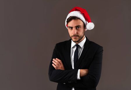 Young handsome caucasian guy in business suit and Santa hats stands on gray background in studio with serious face folded arms on chest Portrait business person with Christmas mood Holiday bannerの写真素材