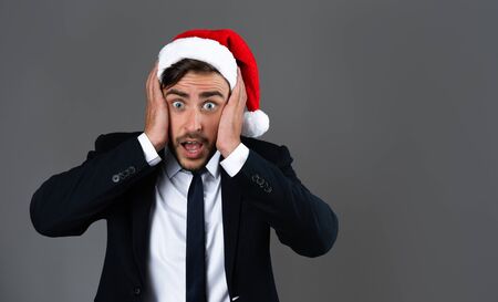 Handsome caucasian man in business suit and Santa hat stands on gray background studio very surprised hands on cheeks Close up portrait business person Fuss concept Holiday banner Forgot to buy gifts.の写真素材
