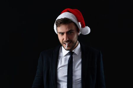 Young handsome caucasian guy in business suit and Santa hat stands on gray background in studio with serious face and smoking cigaretteの写真素材