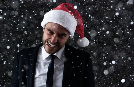 Young handsome caucasian guy in business suit and Santa hats stands on gray background in studio with serious face and smoking cigarette Portrait business person with Christmas mood Holiday bannerの写真素材